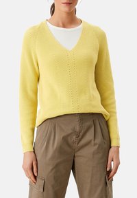 Gelbes Strickpullover mit V-Ausschnitt, gerippter Textur und vertikalen Musterdetails; kombiniert mit hellbraunen Cargo-Hosen.