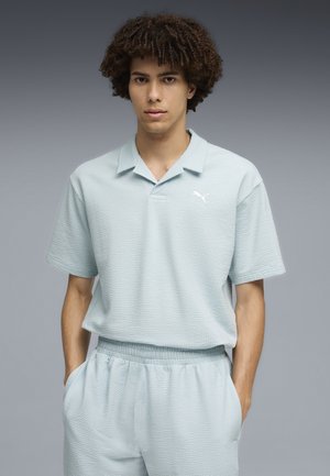 Ung mand med krøllet hår iført en lyseblå, struktureret polo og matchende shorts, står mod en grå baggrund.