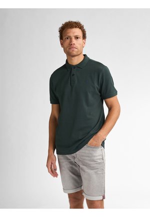 Camisa polo de color verde oscuro con cuello, mangas cortas y tapeta de tres botones, combinada con pantalones cortos claros de color gris, arremangados.