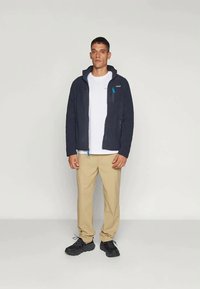 Ναυτικό fleece τζάκετ με φερμουάρ πορτοφόλι, σε αντίθεση γαλάζιες λεπτομέρειες, φορεμένο πάνω από άσπρη μπλούζα, συνδυασμένο με μπεζ παντελόνι και μαύρα αθλητικά παπούτσια.