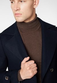 Granatowy wełniany płaszcz z szerokimi klapami, zapinany na podwójne guziki oraz metalowe guziki, nałożony na dopasowany brązowy sweter z golfem.