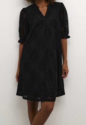 Robe noire longueur genoux avec motif en dentelle florale, manches courtes bouffantes se terminant par des poignets à volants, et un décolleté en V portée par une personne debout.