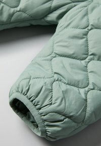 Hiha takki, joka on tehty pehmeästä mintunvihreästä kankaasta. Varustettu joustavalla hihansuulla ja timanttimuotoisella kirjonnalla, joka tuo tekstiiliin tekstuuria.