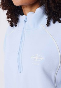 Sweat-shirt zippé bleu clair avec un col haut, des détails en piping blanc et un petit logo sur la poitrine. Texture de tissu douce.