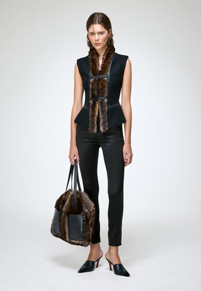 Femme portant un haut sans manches noir avec une bordure en fourrure marron, un pantalon noir, des talons noirs, et tenant un grand sac à main en fourrure noire et marron.