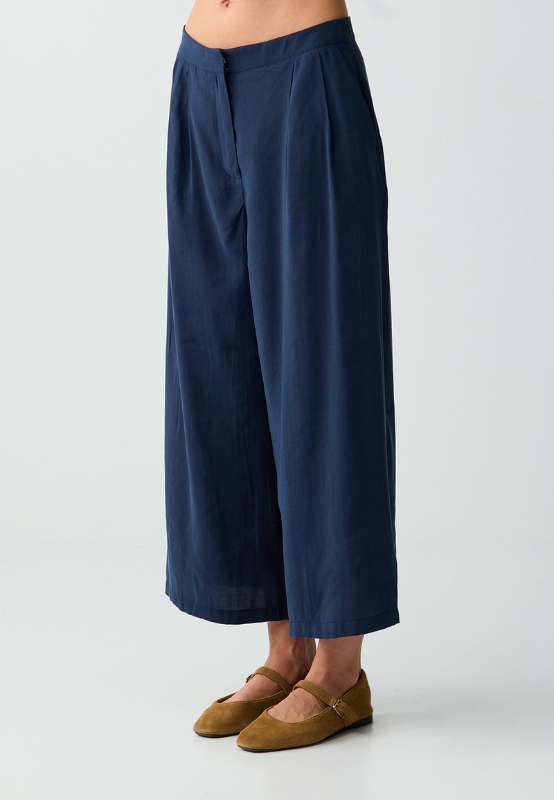 Femme portant un pantalon large bleu marine raccourci et des ballerines Mary Jane en daim marron, debout sur un fond clair uni.