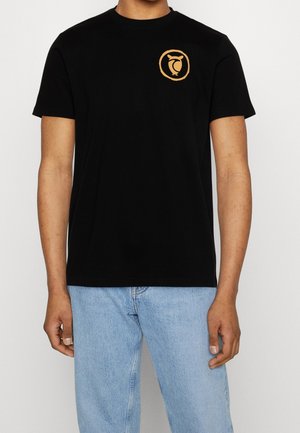 Man draagt een zwart T-shirt met korte mouwen met een klein oranje uilenlogo op de borst en lichtblauwe jeans, staand tegen een witte achtergrond.