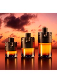 Drie amberkleurige Azzaro parfumflessen in kleine, middelgrote en grote maten, tegen een warme zonsonderganglucht met donkere wolken.