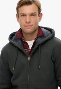 Sudadera gris con cremallera y capucha forrada de forro polar, con una camisa de cuadros roja y negra debajo. Se destacan la cremallera dorada y los cordones.