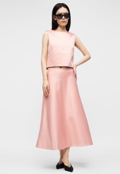 Γυναίκα με μαύρα γυαλιά ηλίου, ροζ τοπ χωρίς μανίκια, φούστα σε σχήμα Α με ψηλή μέση που ταιριάζει και μαύρα μυτερά παπούτσια, στέκεται με το ένα χέρι στη μέση.