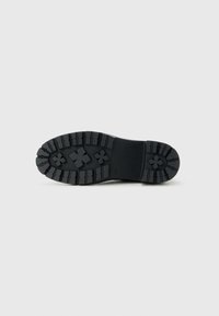 Semelle de chaussure noire avec des rainures profondes et des motifs en forme de croix surélevés, présentée à plat sur un fond gris clair.