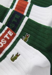 Tejido de punto a rayas verde, blanco y naranja con textura acanalada. Presenta el logo de Lacoste bordado en verde en la sección blanca.