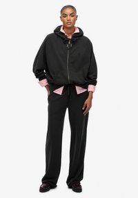 Felpa nera con zip e cordino, indossata sopra una camicia colletto di colore rosa chiaro. Pantaloni neri a taglio rilassato e scarpe scure. Texture liscia.