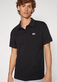 Camisa polo negra en una tela texturizada con cuello tipo solapa. Presenta un pequeño logo de Tommy Jeans bordado en el lado izquierdo del pecho.