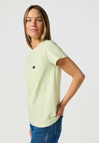 T-shirt di cotone verde chiaro con maniche corte, scollo rotondo e un piccolo logo del marchio blu navy sul petto. Indossato con jeans blu.