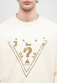 Cremefarget hettegenser med raglanermer, med et slitt gullgrafisk design i trekantform og tekst som sier "GUESS."