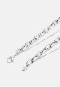 Collier en chaîne argentée avec de grands maillons, une chaîne plus petite, un fermoir en forme de homard, et un pendentif triangulaire avec le nom de la marque "GUESS."