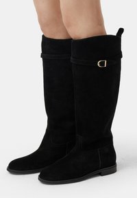 Bottes en daim noires montantes avec un bout rond, une semelle plate et un revers ornés d'une boucle dorée.