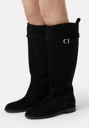 Black suede knee-high boots with a rounded toe, flat sole, and a fold-over cuff featuring a gold-tone buckle detail.