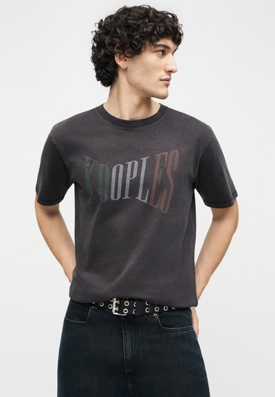 Νέος άνδρας με σγουρά μαλλιά, που φοράει ξεθωριασμένο μαύρο T-shirt με λογότυπο "THE KOOPLES" και μαύρο τζιν με μαύρη ζώνη.