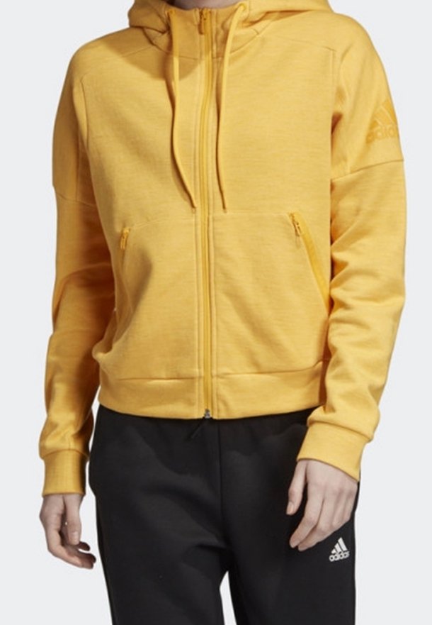 Žlutá mikina na zip s dvěma předními kapsami, měkká tkanina, žebrované manžety a logo na levém rameni. Kombinováno s černými kalhotami.