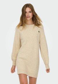 Beige gebreide jurk met lange mouwen, ronde hals en drie gouden knopen op de linkerschouder. Zachte textuur, midden boven de knie.