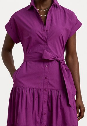 Frau trägt ein kurzärmeliges violettes Hemdkleid mit gebundenem Taillengürtel und geschichtetem Rock, Hände in den Taschen und Muschel-Halskette.