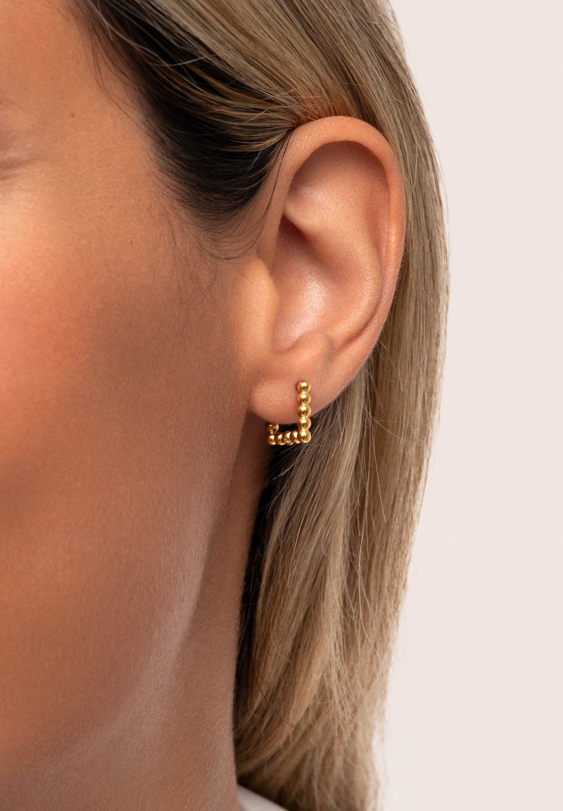 Orecchino a cerchio in oro con una texture a perline, caratterizzato da un design geometrico e piccole sfere lungo la superficie, esibito su un tono di pelle chiara.