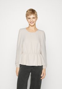 Light beige long-sleeve top with a rounded neckline and peplum waist. Soft, textured fabric with gathered sleeves, styled with dark gray pants.