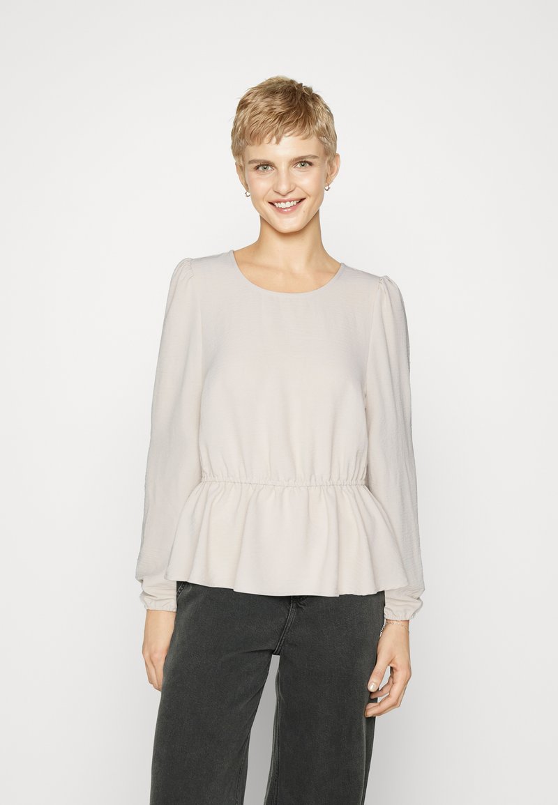 Light beige long-sleeve top with a rounded neckline and peplum waist. Soft, textured fabric with gathered sleeves, styled with dark gray pants.