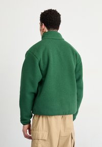 Vihreä fleece-takki, jossa on korkea kaulus, teksturoitu kangas ja rento istuvuus, yhdistettynä beigeihin cargo-housuihin.