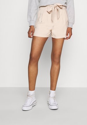 Person iført beige højtaljede shorts med bundet bælte, grå sweatshirt, hvide sokker og hvide høje sneakers stående mod en hvid baggrund.