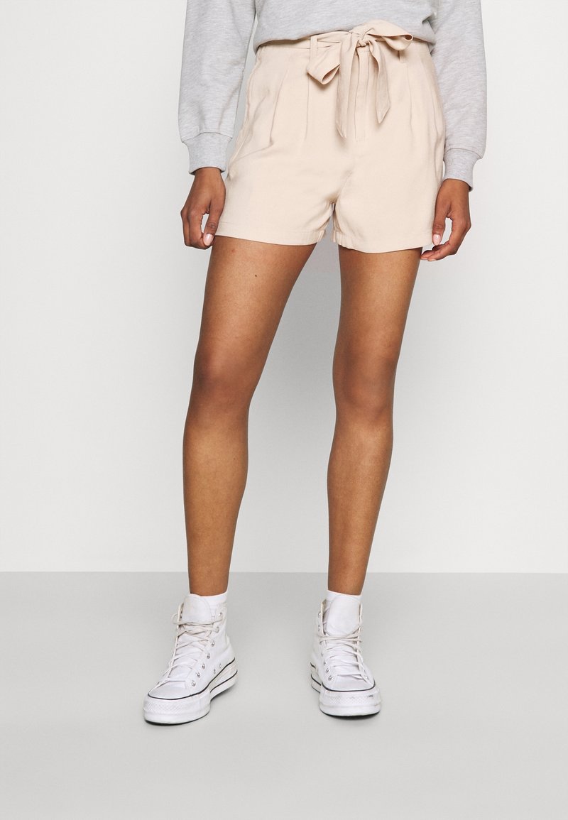 Personne portant un short beige taille haute avec une ceinture nouée, un sweat gris, des chaussettes blanches et des baskets montantes blanches, debout devant un fond blanc.