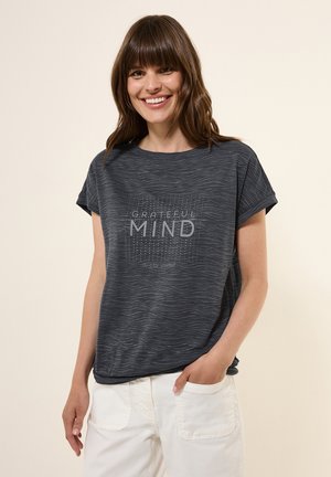 Dark gray short-sleeved t-shirt with a wave pattern. Centered printed text reads "GRATEFUL MIND" with a grid design. Paired with light-colored pants.