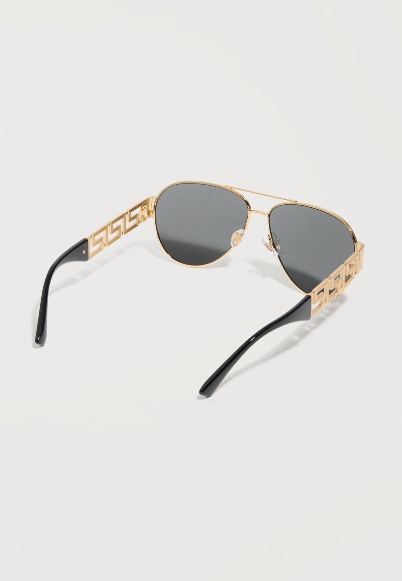 Lunettes de soleil aviateur à monture dorée avec verres foncés et embouts de branches noirs, présentant un motif découpé géométrique sur les branches.