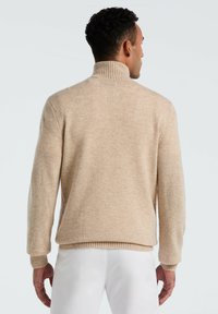 Mann, der sich abwendet, trägt einen beigen Rollkragenpullover und weiße Hose vor einem schlichten hellen Hintergrund.