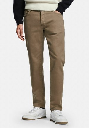 Man die beige rechte broek en witte sneakers draagt, staat met één hand in de zak tegen een effen achtergrond.