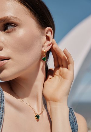 Des boucles d'oreilles et un collier en ton doré ornés de cristaux verts en forme de cœur. Le modèle présente ces pièces sur fond d'un ciel bleu vif.
