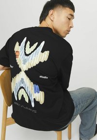 Sort T-shirt med korte ærmer, der har et farverigt abstrakt design på ryggen i nuancer af blå, grøn og beige, med hvide tekstaccenter.