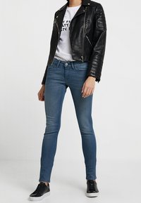 Giacca da motociclista nera in pelle con dettagli a coste, t-shirt bianca con grafica, jeans slim-fit blu e sneaker nere con suola bianca.