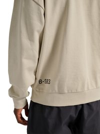 Personne portant un sweat-shirt beige à manches longues avec « B-583 » imprimé près de l'ourlet inférieur et un pantalon noir, vue de dos.