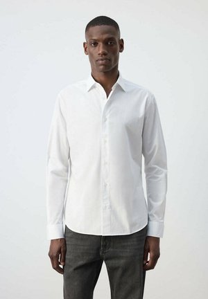Hombre de pie frente a un fondo liso, vistiendo una camisa blanca de manga larga con botones y pantalones gris oscuro, mirando hacia adelante con los brazos relajados.