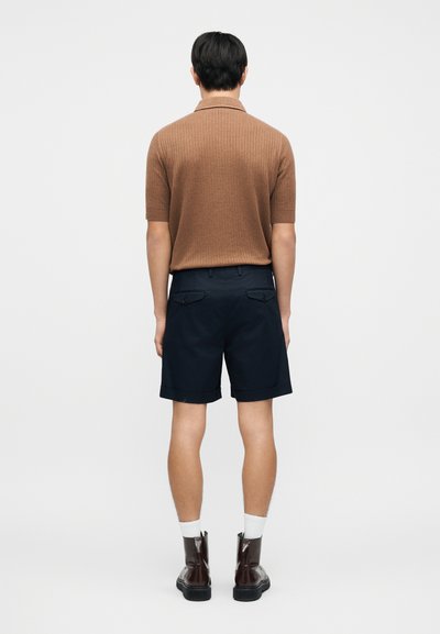 Καφέ ριμπ μπλουζάκι πόλο με κοντά μανίκια συνδυασμένο με σκούρες μπλε σορτς. Το μοντέλο φορά καφέ μποτάκια και λευκές κάλτσες, με ορατές λεπτομέρειες πίσω τσέπης στα σορτς.