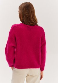 Różowy, puszysty sweter z luźnym krojem, okrągłym dekoltem i długimi rękawami. Gładka faktura, lekko skrócony dół. Noszony z jasnymi spodniami.