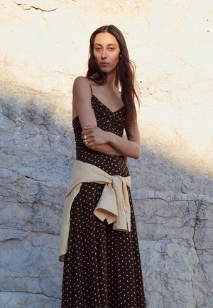 Kobieta z długimi, ciemnymi włosami w brązowej sukience w grochy, z kremowym swetrem zawiązanym w talii, stojąca na tle fakturowanej kamiennej ściany w świetle słonecznym.