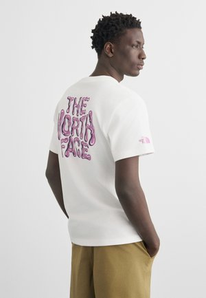 Giovane uomo con capelli corti e ricci, che indossa una maglietta bianca con la scritta viola "The North Face" e pantaloni beige, in piedi con le mani nelle tasche.