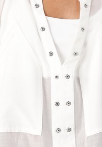 Top bianco con scollatura a V, caratterizzato da bottoni esagonali in argento e tessuto testurizzato. I dettagli degni di nota includono accenti cuciti e un design a strati.