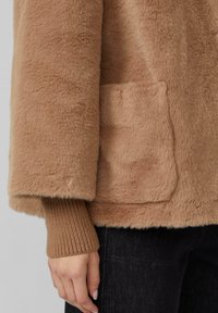 Veste beige duveteuse avec une finition texturée, dotée d'une poche latérale et de poignets côtelés marron. Fait partie d'une tenue en denim foncé.