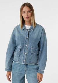 Kobieta w jasnoniebieskiej dżinsowej kurtce z postrzępionym dołem i kieszeniami naszytymi, w połączeniu z dopasowanymi jasnoniebieskimi dżinsami na białym tle.