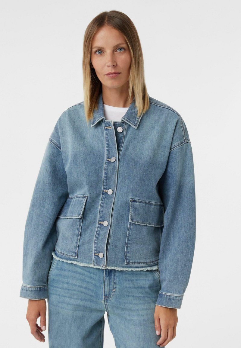 Kobieta w jasnoniebieskiej dżinsowej kurtce z postrzępionym dołem i kieszeniami naszytymi, w połączeniu z dopasowanymi jasnoniebieskimi dżinsami na białym tle.
